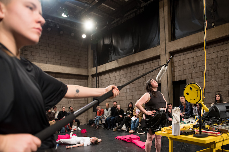 Jen Rosenblit speaks into a boom mic which is held over her head by a performer who wears a black t-shirt. Other performers are in the background laying down with the fuchsia fabric 
